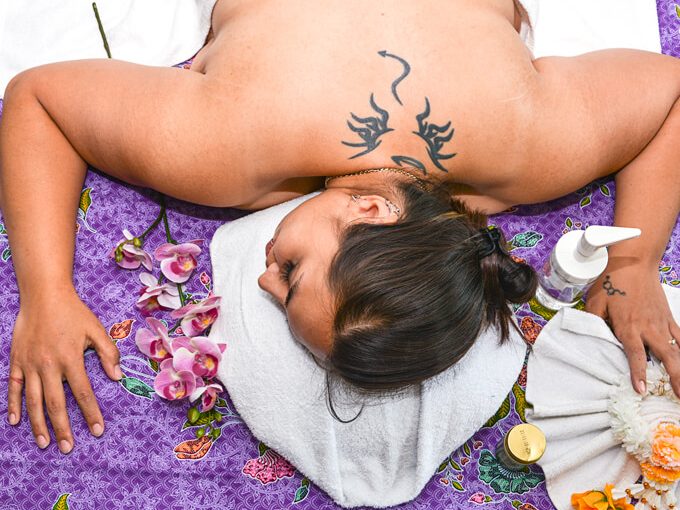 Blick von oben auf eine Aroma-Massage: Kundin auf einer lilafarbenen Matte, daneben Öle und Orchideenblüten.